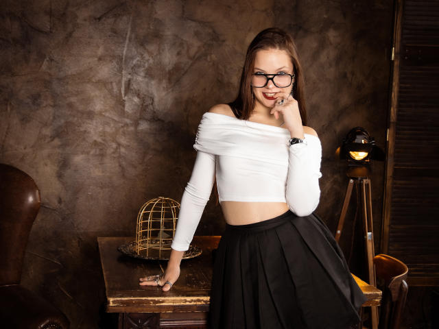 BethCutie com óculos, blusa branca com um ombro descoberto, saia preta, em pé, com a mão na mesa, gaiola de pássaro, luminária de madeira, parede marrom, sorrindo, dedo no queixo, cabelo castanho, fofa..