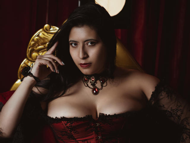 KamileNilsen with large tits, wearing a red and black lace corset, black choker with red gem, and black lace sleeves, looking seductively at the camera. Dark hair, red lips.