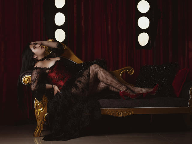 KamileNilsen has black hair, red lipstick, and red heels. She's wearing a black lace dress, red corset, and fishnet stockings, lying on a gold chaise, showing her ass.