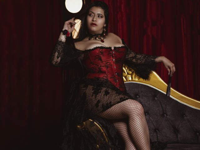 KamileNilsen with big tits in a red-black lace corset, fishnet stockings, and dark makeup, sitting on a gold-grey couch, holding a black cane, against red velvet curtains.