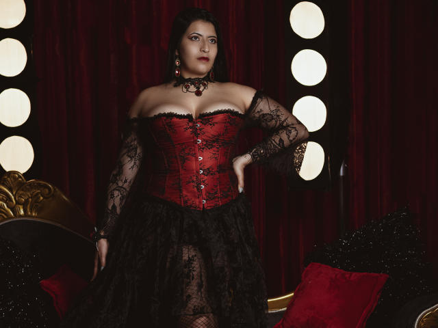 KamileNilsen with large tits in a red lace-up corset, black lace sleeves, and black lace skirt, standing confidently, hands on hips, dark red curtain background, glowing lights.