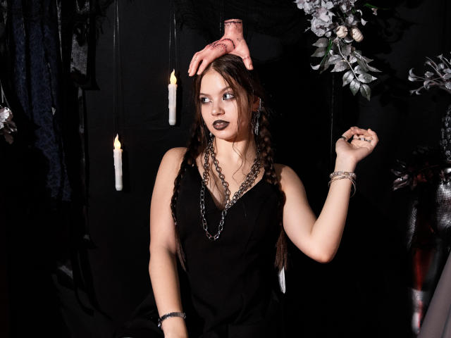 BethCutie stands centered, wearing a black sleeveless dress with a deep V-neck, chains around her neck, and braided hair. A pink hand rests on her head while she looks to the side with dark lipstick. Her right hand raises, showing a ring. Dim, lit candles and dark floral arrangements are in the background. The composition is dramatic and moody, with a strong contrast between BethCutie's pale skin and the dark surroundings.