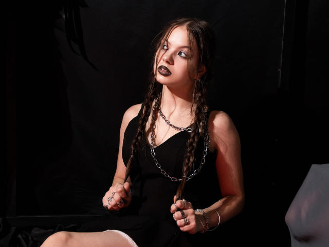 BethCutie mit geflochtenem, braunem Haar, schwarzem Lippenstift, einer Kettenkette, einem schwarzen Kleid, die eine Strähne hält, sitzend, dunkler Hintergrund, nach oben schauend, Metallringe an den Fingern, blasse Haut, leichte Schatten..