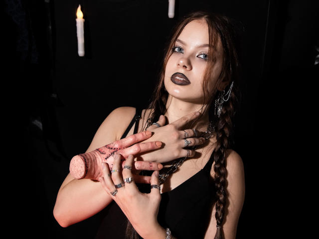 BethCutie, a pale-skinned woman with dark braided hair, stares forward with black lipstick and blue eyes. She wears a black tank top, pink lace fingerless gloves, and multiple rings on her fingers. Her hands are raised, fingers touching her neck. The background is dark with two lit candles. Her makeup and gloves have a gothic, punk style. The lighting highlights her face and hands, creating a dramatic, eerie atmosphere.