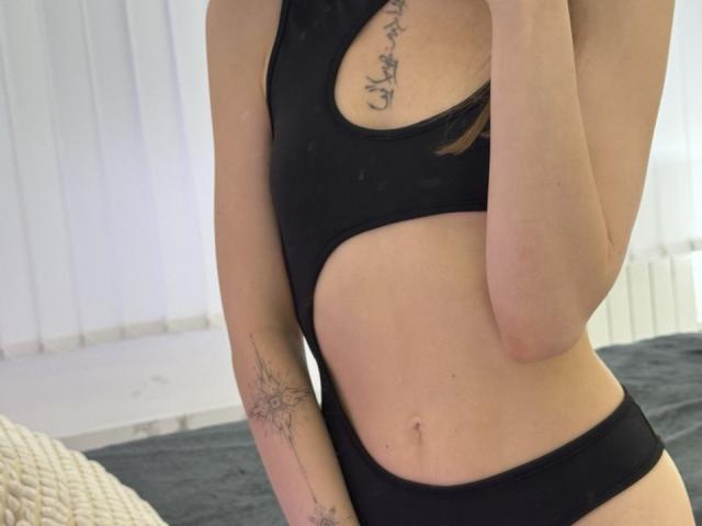 EmiLake draagt een zwart badpak met een uitsparing, waardoor haar slanke taille, kleine borsten en tatoeages op haar borst en arm zichtbaar zijn. Ze houdt haar telefoon vast en bedekt haar gezicht..