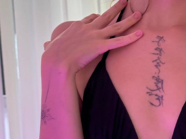 EmiLake heeft een zwarte tatoeage op haar borst en een bloementatoeage op haar arm. Ze draagt een zwarte jurk, met haar hand op haar sleutelbeen..