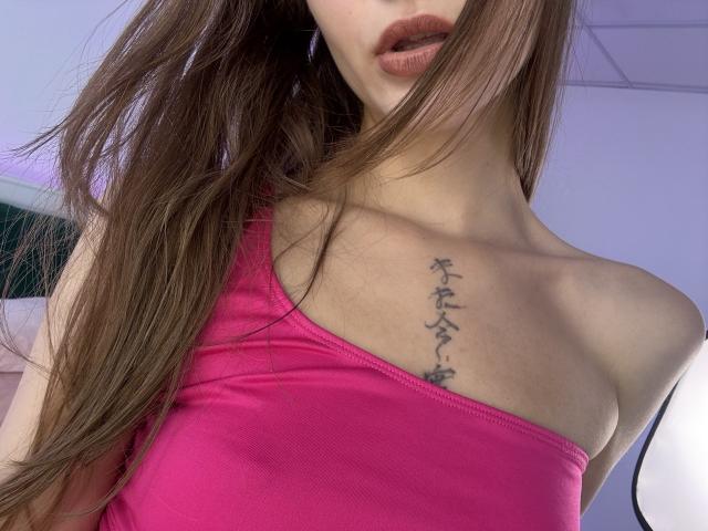 EmiLake with long brown hair, pink tank top, showing off a small tattoo of Japanese characters on her right chest, and her slightly open red lips.