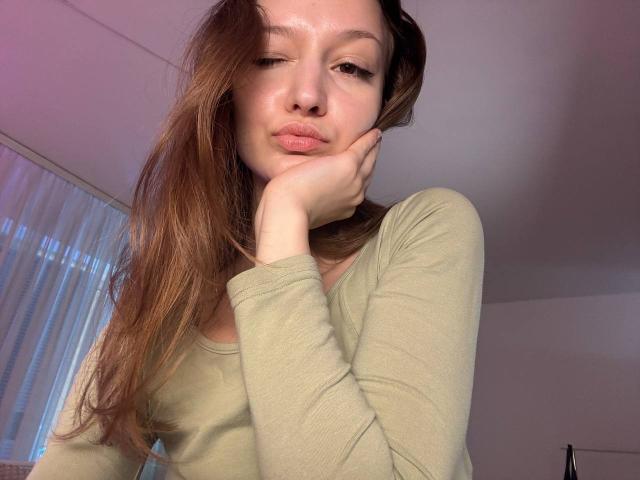 EmiLake with long brown hair, wearing a green long-sleeve shirt, pouting with her hand under her chin, showing a cute, slightly smug expression.