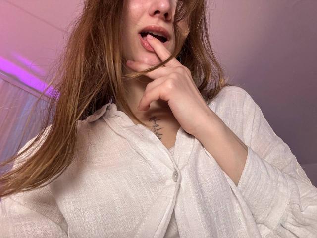 Emi, met lang bruin haar, een wit overhemd met knopen, een vinger in haar mond, een subtiele tatoeage op haar nek. Geen borsten, geen zichtbare billen of vagina..
