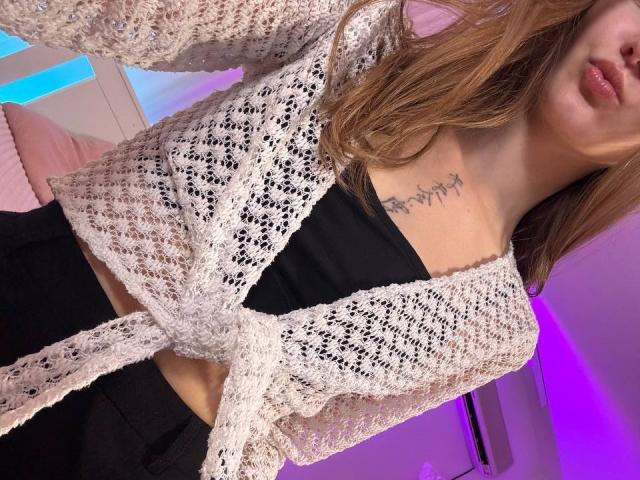 EmiLake with wavy brown hair, wearing a white knit cardigan tied at the front, black tube top showing her tits, black pants, and tattoo under her right boob.
