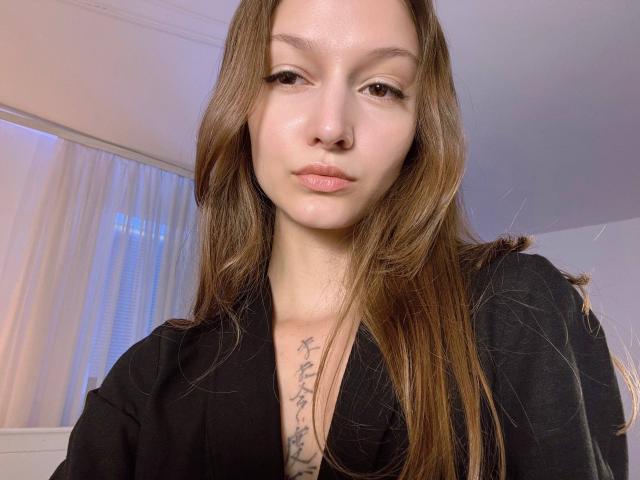 Emi, met lang bruin haar, een zwart shirt, een tatoeage op haar borst, een neusring en subtiele make-up, staart kalmpjes in de camera. Zacht licht op haar gezicht..