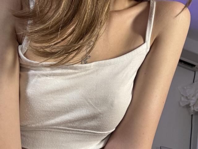 EmiLake's chest showing a white, spaghetti-strap tank top, her small tits slightly visible, with light brown hair covering part of her upper chest. Her skin is fair.