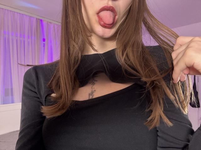 EmiLake with brown hair, black top, and cutout chest, sticking out her tongue, showing a tattoo under her tits. She's holding her hair with her right hand.