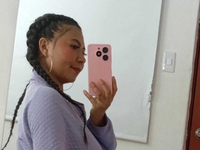 JadeFoxx mit geflochtenen Haaren, die ein lila Hemd trägt, ein rosa Handy hält, lächelt und ihr Profil zeigt, mit großen Reifenohrringen und einem Ring, in einem Spiegel-Selfie..
