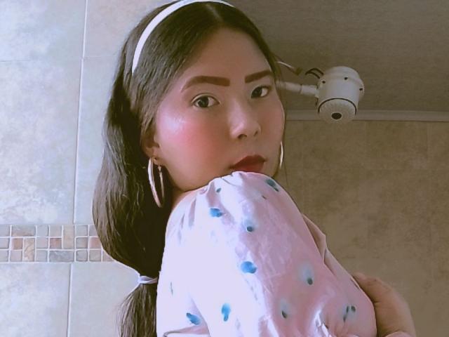 JadeFoxx trägt rosigen Lippenstift, einen weißen Haarreif und ein rosa Hemd mit blauen Punkten. Sie hält sich am Arm, blickt zurück mit rosigen Wangen und trägt große Ohrringe. Sie befindet sich in einem Badezimmer..