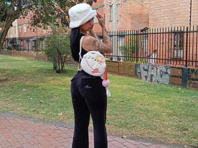 MadisonDarkk stands with her back to the camera, wearing a white furry hat, black velvet pants, and a small white stuffed cat bag with pink ears and a red ribbon. She has long black hair, a tattoo on her left arm, and a curvy, hourglass figure. The background includes a brick building, black iron fence, and graffiti on a wooden wall. The ground is a mix of green grass and red brick pavement. She looks to the right.