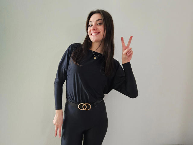MiaLou-hot stands against a plain background, smiling with dark, straight hair. She wears a black long-sleeve top and black pants with a gold belt featuring a double loop design. Her right hand is raised, making a peace sign with her fingers. She has a small gold necklace. The image is clear, with a simple composition focusing on MiaLou-hot. Her relaxed, confident pose is highlighted by her smile.