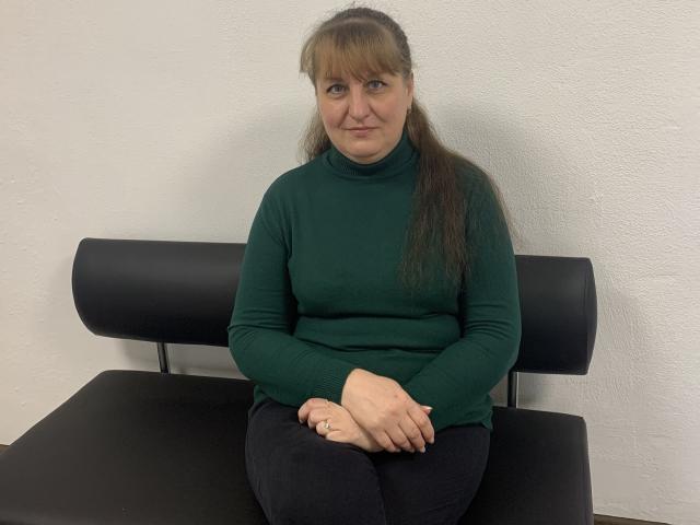 JessicaMery with long brown hair and bangs, wearing a green turtleneck, black pants, and sitting on a black chair with hands folded, looking slightly to the right.