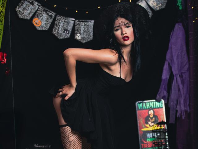 LizzeJoy with black dress, fishnet stockings, and huge fluffy black hat, bending over with hand on ass, red lipstick, and black lace over her eyes. Dark, sexy, and seductive.