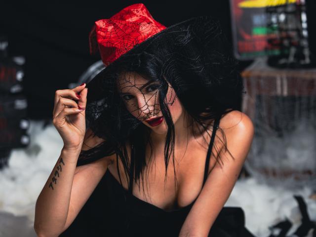 LizzJoy with black hair, red hat, web-like face covering, and deep cleavage, touching her hat, showing tattoo on her right arm, and red lipstick. Boobs visible, sexy look.