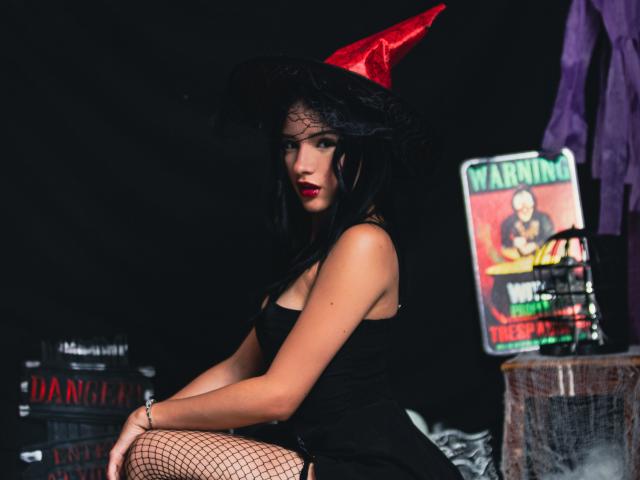 LizzJoy with black dress, fishnet stockings, red hat, and red lipstick, sitting on a rock, showing her ass, in a dark, gothy setting with a warning sign.