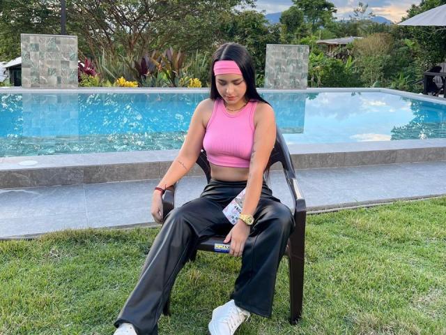 ZoeCastelli sitting on a black chair by a pool, wearing a pink sports bra, black pants, white sneakers, pink headband, and a wristwatch, with her ass slightly visible.