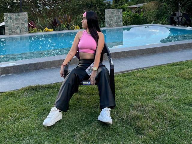 ZoeCastelli with long black hair, pink sports bra, black leather pants, white sneakers, sitting on black chair, pool behind, grass in front, looking left, holding phone.