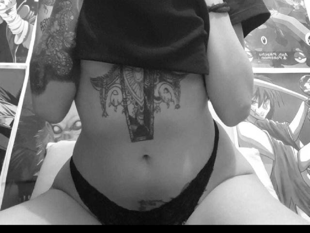 HaryBlair's bare belly, showing a detailed tattoo just below the tits, wearing black lace panties, lifting a shirt, with tattoos on the right arm, in a black-and-white photo.