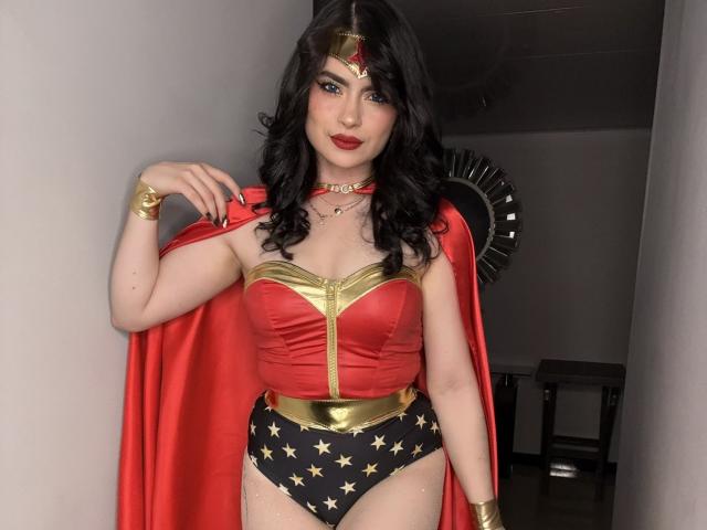 ClariceMontclair with black wavy hair, red lipstick, wearing a Wonder Woman costume: red corset, gold bust, black star-patterned panties, red cape, gold tiara, and bracelets, smiling.
