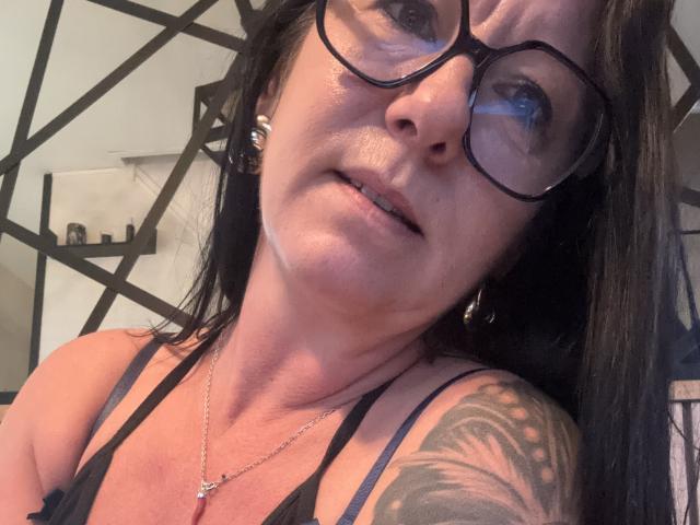 CoquineX69 with glasses, black hair, slight smile, tattoo on right shoulder, necklace, black tank top, showing her tits, ear piercings, looking up, slightly messy skin, indoor lighting.
