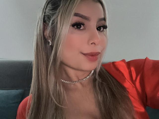 AmaraRicci has long blonde hair, thick eyebrows, and is wearing a red shirt with a pearl choker. She's looking at the camera with a slight smile.