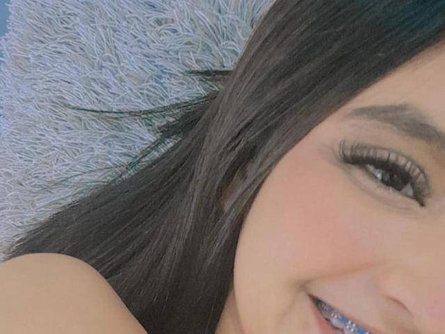 Mharianita's face with long black hair, dark eyebrows, and big eyelashes, smiling slightly with her upper chest visible, lying on a fluffy gray pillow.
