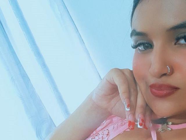 Mharianita with pink lace bra, nose ring, and red lipstick, showing off her painted nails and gently resting her chin on her hand. Her tits are partially visible.