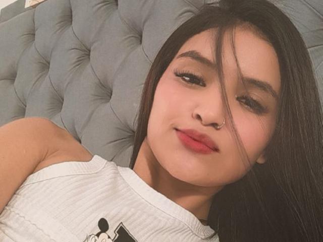 Mharianita with long black hair, nose ring, and red lipstick, lying on a tufted grey headboard, wearing a white ribbed tank top, smiling slightly.