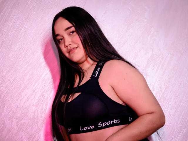 EmilyBrice with long black hair, wearing a black Love Sports bra, showing her tits, smiling slightly, against a pink-lit white wall. She has a cute, relaxed expression.