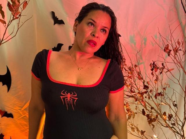 LizethBells, a Latina woman with wet dark hair, wears a tight black shirt with red trim and a spider emblem, red lipstick, and a gold necklace. She stands against a backdrop with bats and dry branches, lit by red and orange light. Her expression is neutral, slightly curious. The composition focuses on her upper body and face.