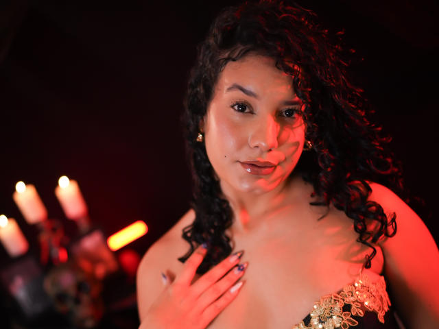 RachellBeaumont with curly black hair, light brown skin, and glossy lips, covering her tits with a hand, wearing a gold-embellished strapless top, surrounded by red candlelight.