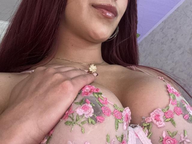AnyaJay with dark red hair, glossy pink lips, wearing a pink floral lace bra, holding a small flower against her large tits, gold necklace, and hoop earrings.
