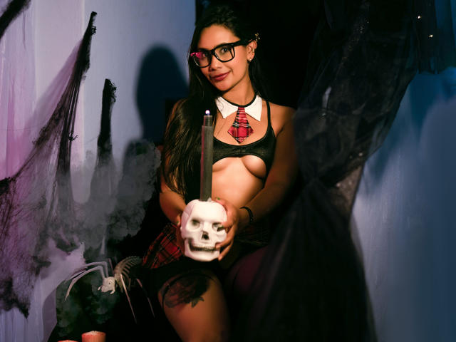 SophieJones69 with black glasses, red tie, and black top, holding a skull, showing her tits, dark hair, and a tattoo on her thigh, in a spooky, dark setting.