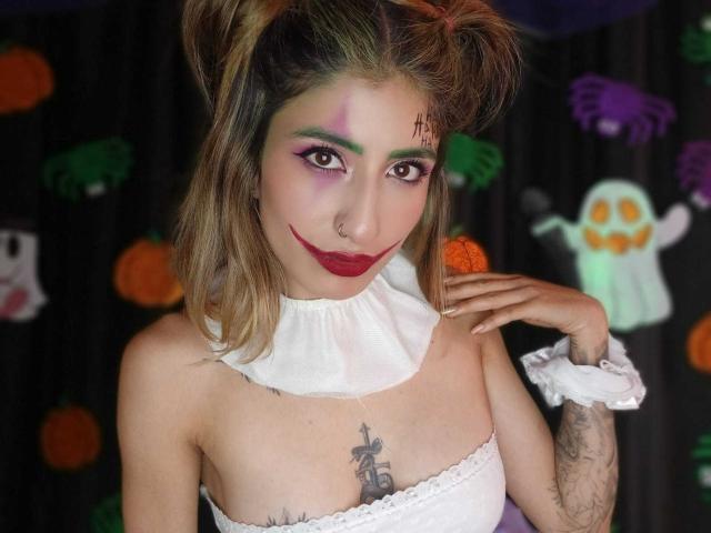 Kaiix with green and purple eye makeup, red lipstick, white ruffled top, and puffy brown hair, showing tattoos and nose ring, with hand near face, smiling.