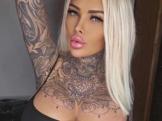 MilaElaine-hot with platinum blonde hair, full pink lips, nose ring, tattoos on neck and chest, and black top, showing her tits and arm tattoos.