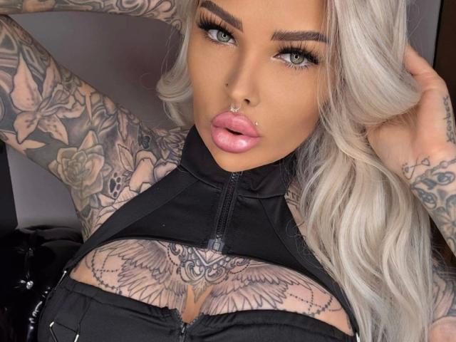 Tattooed MilaElaine-hot with platinum blonde hair, full pink lips, nose ring, and eyeliner, wearing a black halter top, exposing her tits with a wing tattoo.