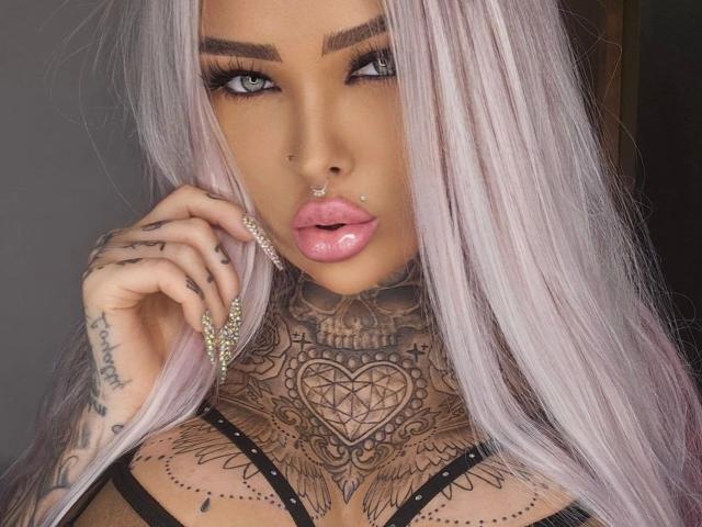 MilaElaine-hot with pink hair, tattoos, nose ring, lip piercings, and full pink lips, holding a chain, showing off her chest tattoos. Boobs and hand tattoos visible.