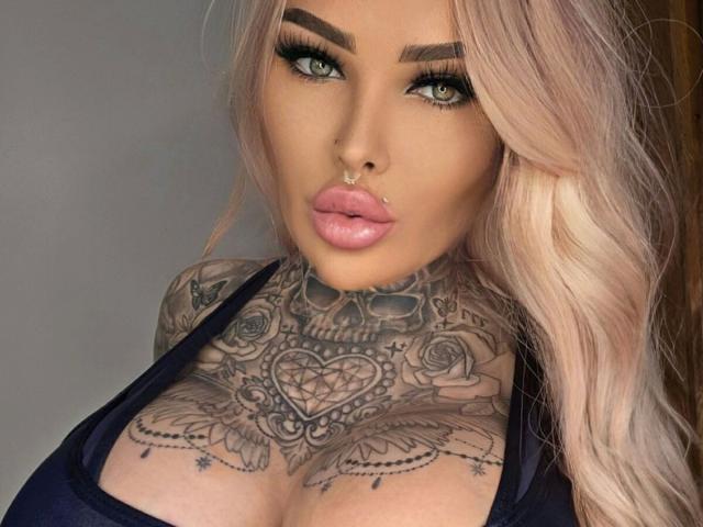 MilaElaine-hot has platinum blonde hair, green eyes, and a nose ring. Her tattoos cover her chest and neck, including a heart and rose design. She's wearing a dark top.