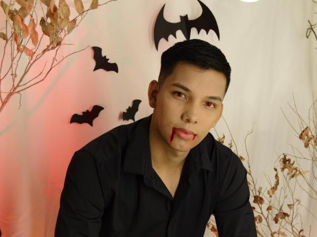 EdwinBlaze with bloody nose, black shirt, short black hair, and bat decorations, looking at camera, with red and white lighting, and dried branches in the background.