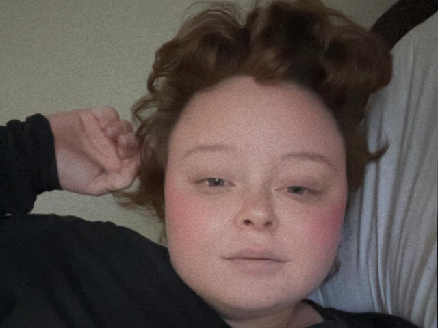 BirdoBih with curly brown hair, light skin, and pink cheeks, lying down with a black shirt, slightly smiling, right hand near head, looking relaxed.