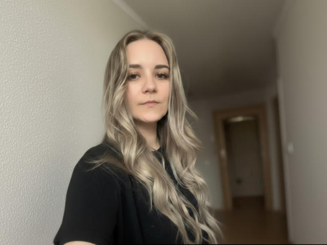 CandiCage with long, wavy blonde hair, wearing a black shirt, standing in a hallway, looking slightly to the side with a neutral expression.