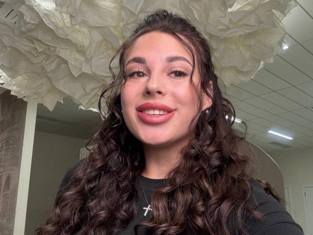 BlackFatal, com cabelo castanho encaracolado, batom rosa e um colar com uma cruz, sorrindo para a câmera, vestindo uma camisa preta, com um teto branco que se assemelha a papel acima..