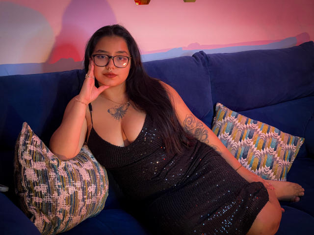 SofiDollg with long black hair, glasses, and a sparkly black dress, showing her tits and a butterfly tattoo, lounging on a blue couch with patterned pillows.