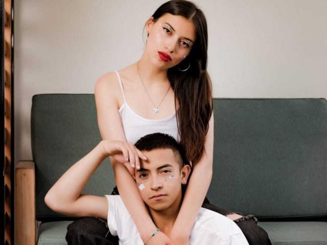 CrimsonSecretts, a chick with red lipstick, white tank top, and long brown hair, sits on a dude's lap, her hands on his face, his hands on her thigh.
